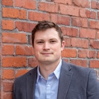 George Sanderson has short dark hair and is standing in front of a brick wall